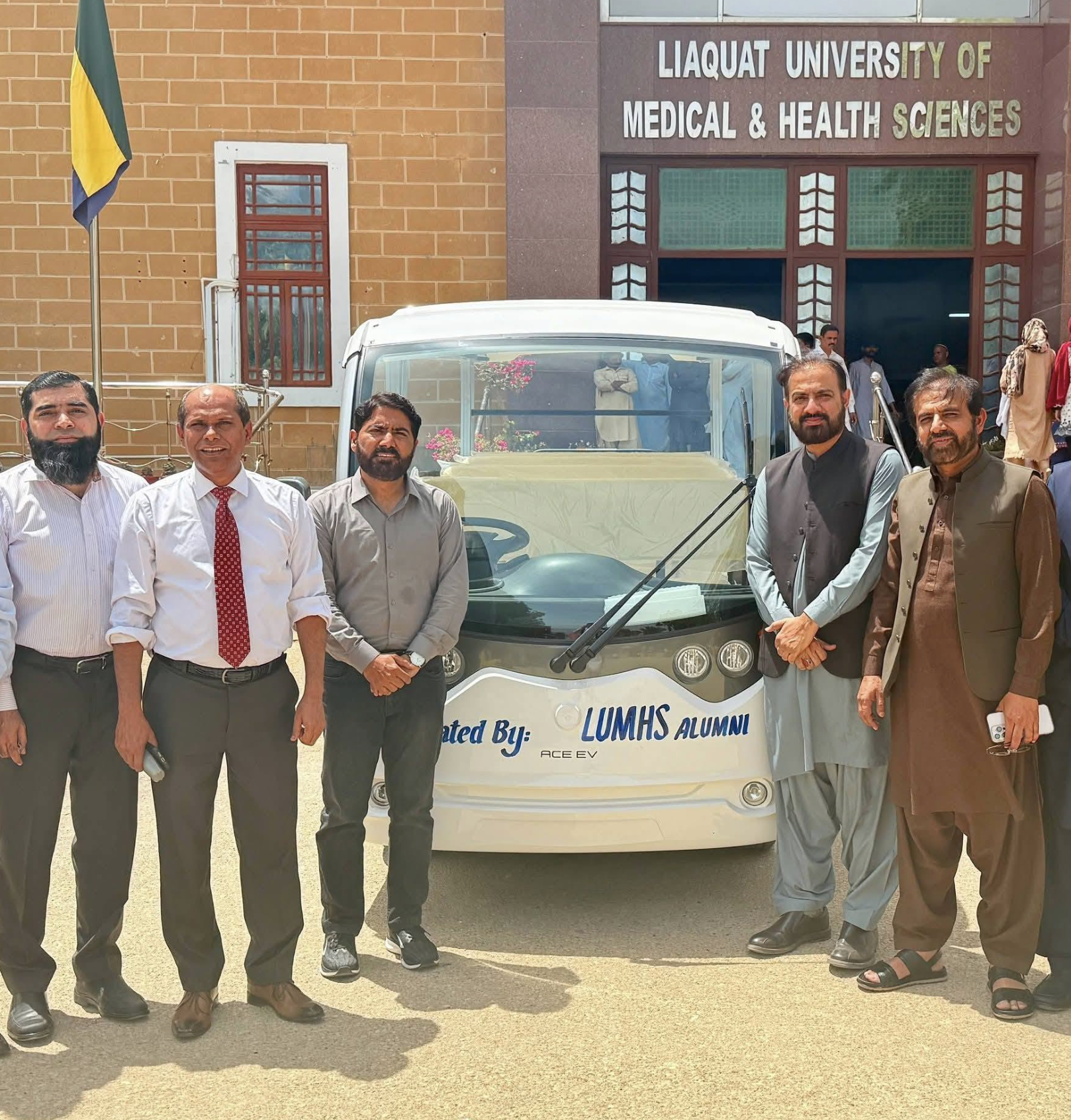A group standing with the electric shuttle outside Liaquat University of Medical and Health Sciences; the vehicle bears LUMHS Alumni lettering.